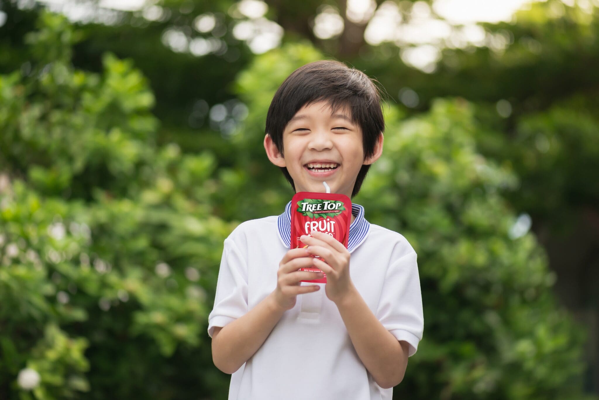 Smiling child outdoors, holding a red Tree Top fruit juice pouch toward the camera.