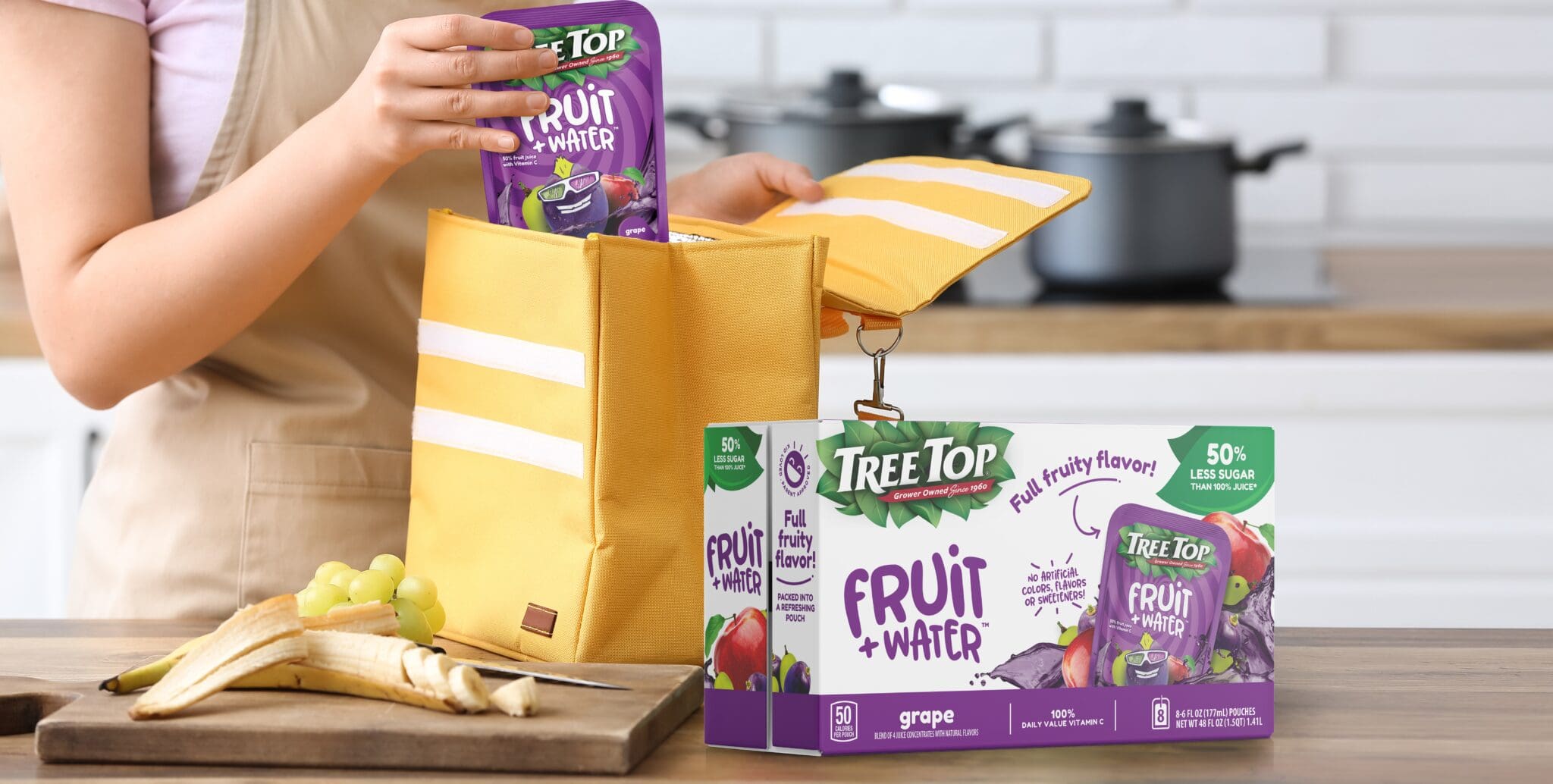 Person places a purple Fruit + Water pouch into a yellow tote bag on a kitchen counter, with a box of Tree Top Fruit + Water drinks nearby.