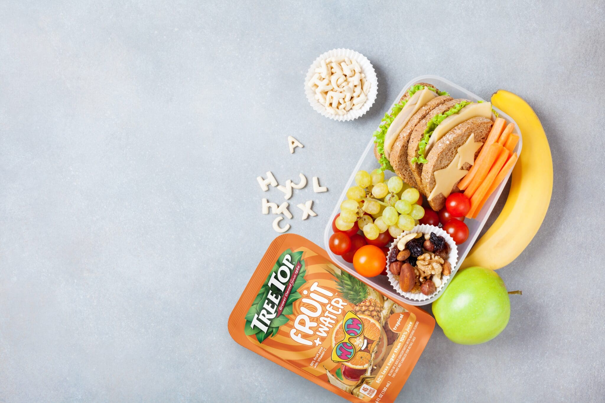 Overhead view of a snack tray with bread slices, cheese, lettuce, carrot sticks, grapes, tomatoes, and a banana, on a gray surface