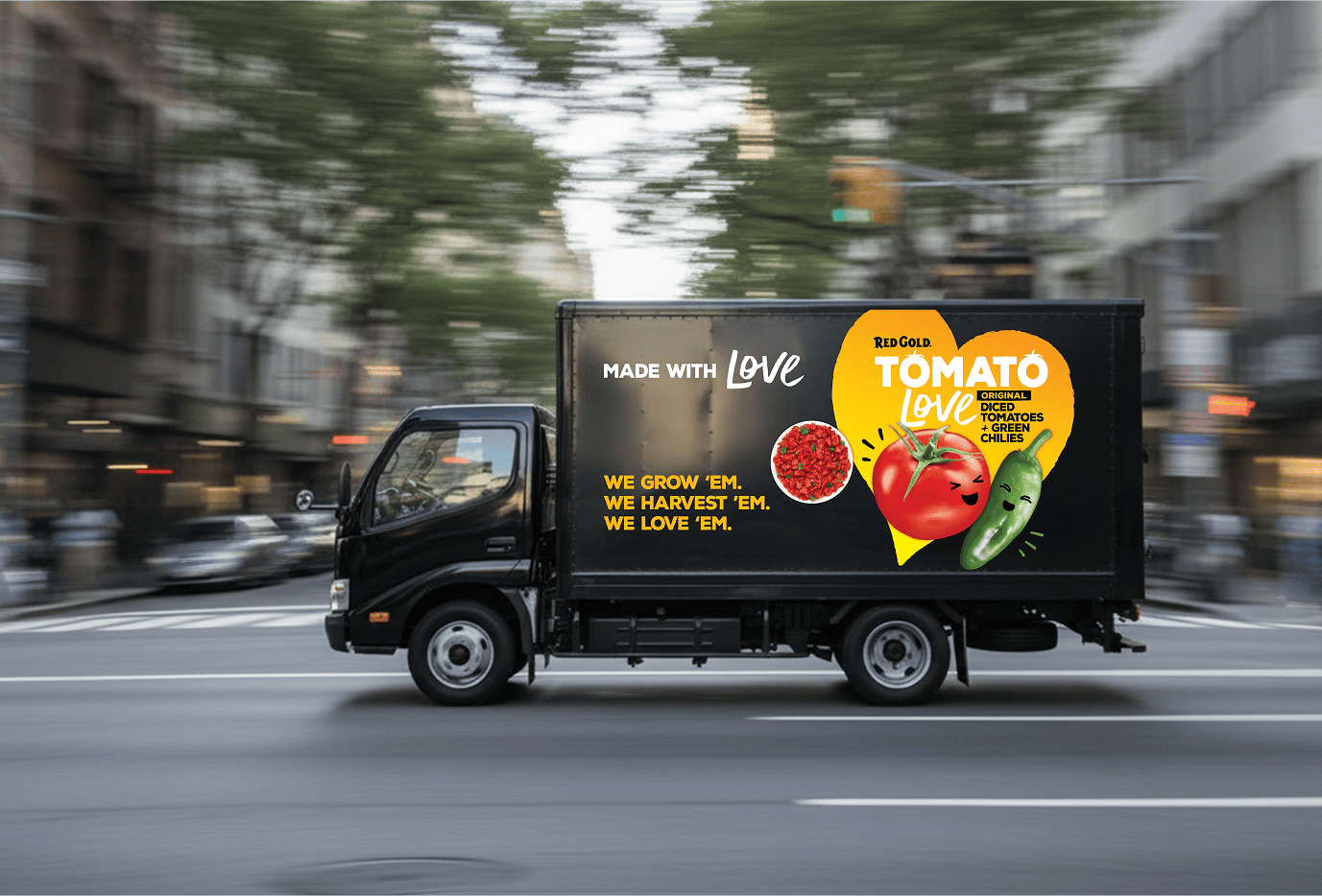 Delivery truck speeding through a city street with a black advertisement panel showing the Red Gold Tomato Love logo and smiling tomato and peppers illustration, plus text 'Made with Love' and 'We grow ’em. We harvest ’em. We love ’em.'