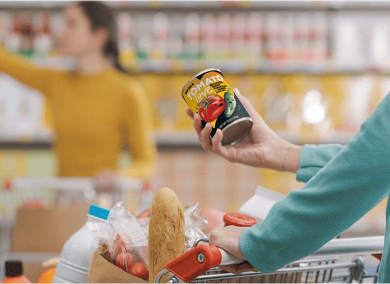 Shopper selecting a can of tomato sauce while pushing a grocery cart in a supermarket. A loaf of bread and other groceries are visible in the cart.