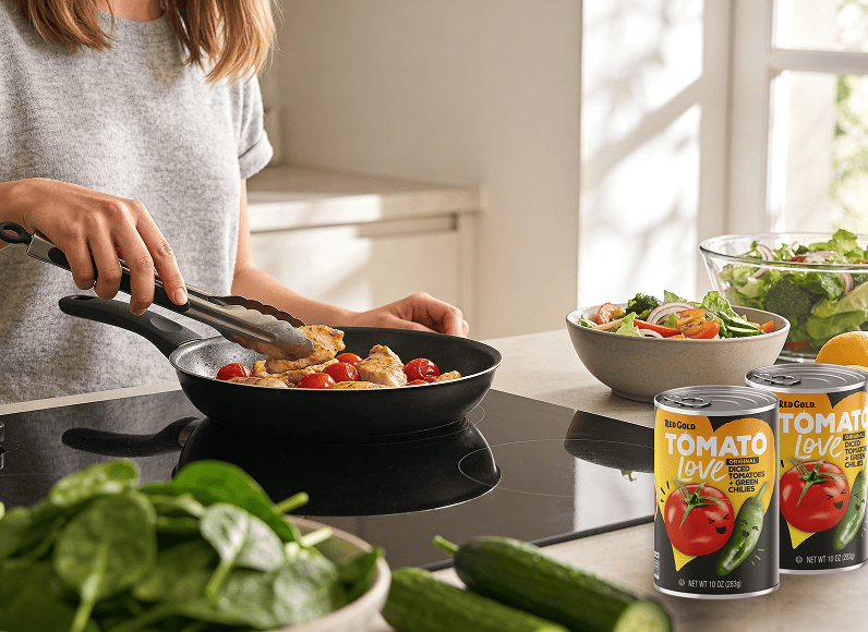 Person cooking chicken and cherry tomatoes in a skillet on the stove, with a salad and tomato cans nearby in a bright kitchen.