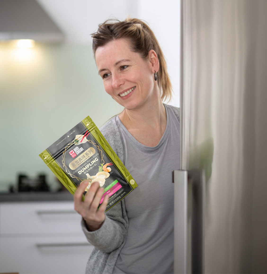 Smiling woman holding a dumpling mix packet near a stainless steel fridge in a kitchen.