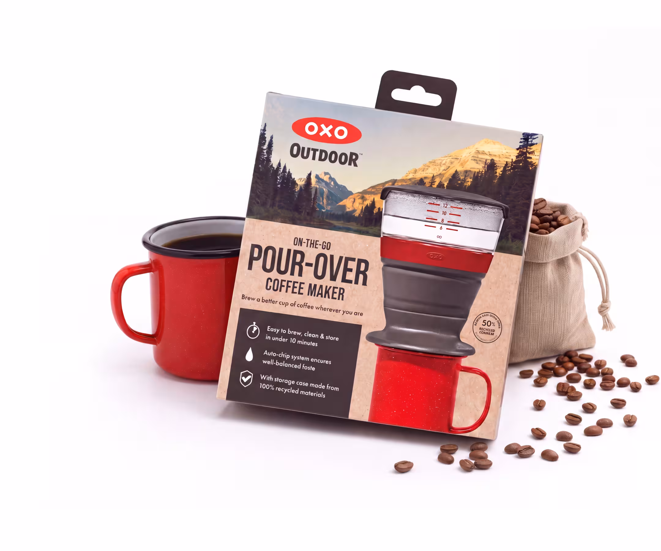 Package for OXO Outdoor Pour-Over Coffee Maker leaning on a red mug with scattered coffee beans nearby, outdoorsy mountain-themed packaging visible.
