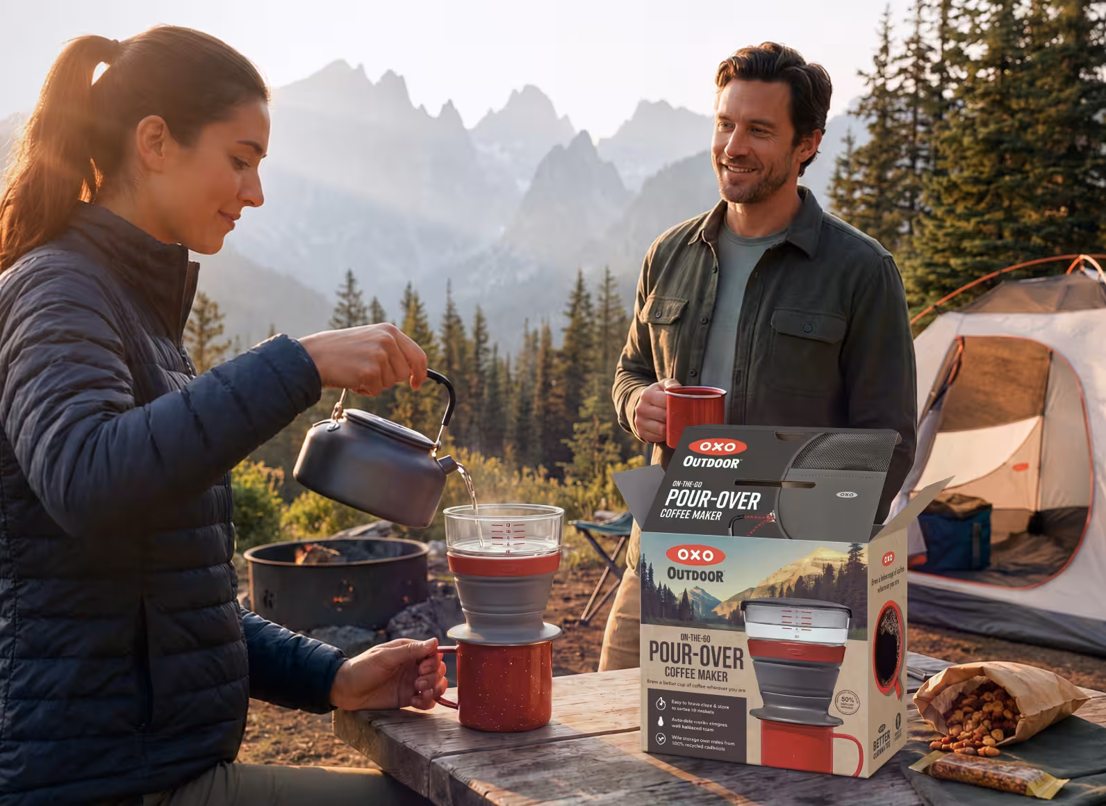 Two hikers camped in a mountain clearing; a woman pours hot water into a red pour-over coffee maker on a wooden table while a man holds a mug and smiles, with a tent in the background.