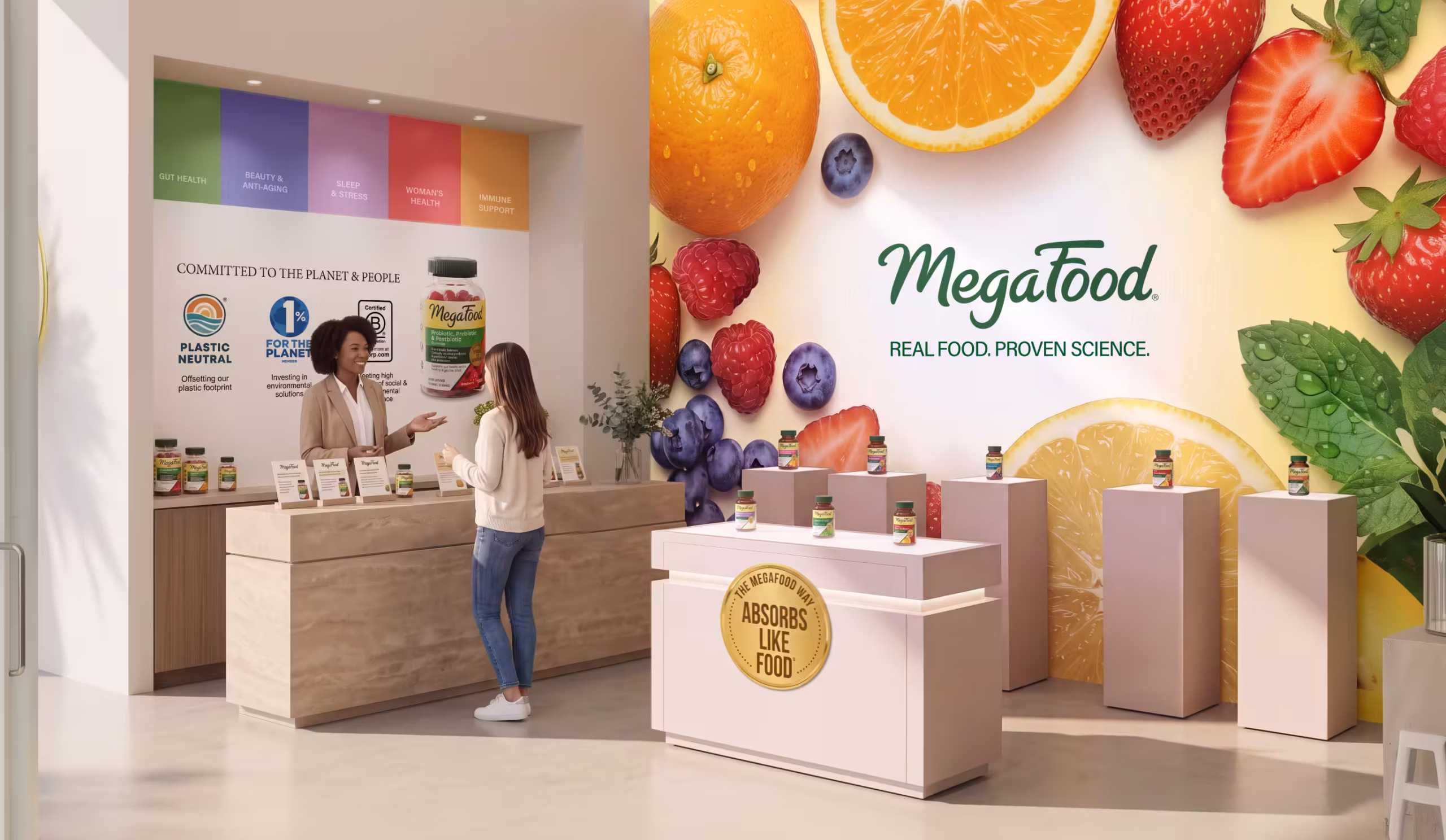 Two women talk at a Megafood booth with fruit-themed wall and product display bottles on a wooden counter and pedestal stands.