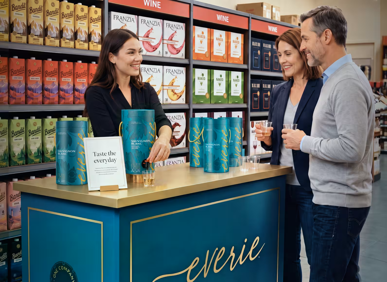 Sales associate pours wine samples for a couple at a teal tasting counter in a wine shop, with colorful boxed wines on the shelves behind them.