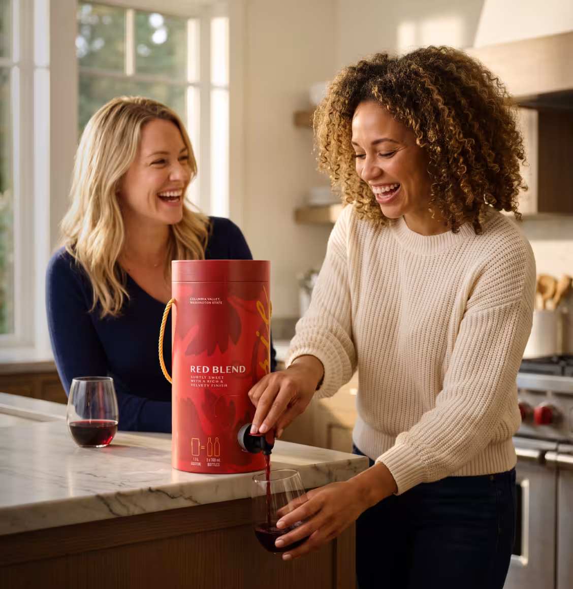 Two women in a bright kitchen laugh as one pours red wine from a red cylindrical container into a glass on a marble countertop.